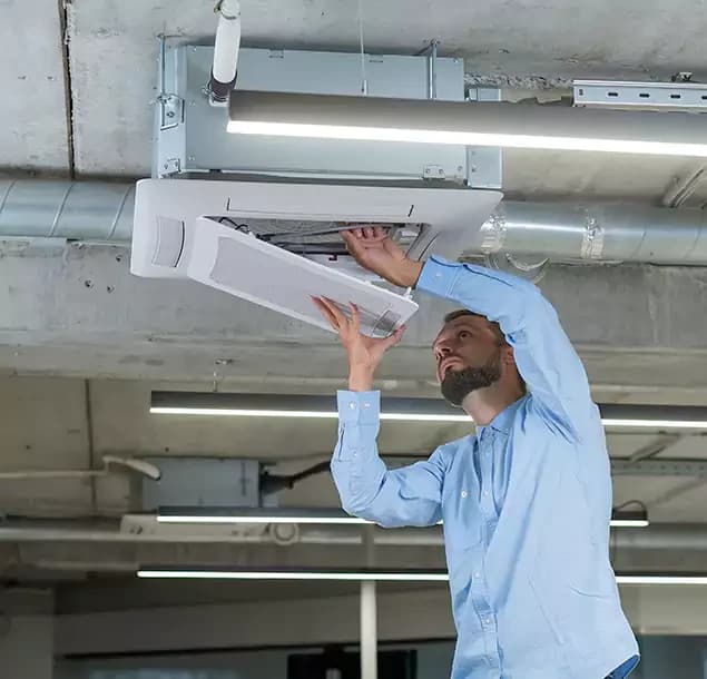 A technician servicing a ceiling cassette air-conditioning unit beneath exposed ductwork and lighting in a commercial interior